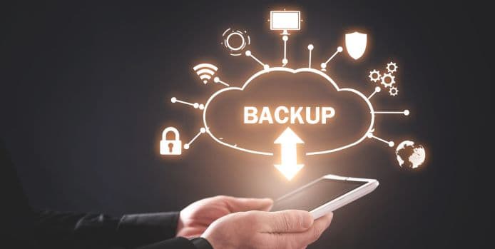 Hands holding a tablet with a glowing cloud icon labeled 'BACKUP,' surrounded by technology symbols representing data storage, security, and connectivity.